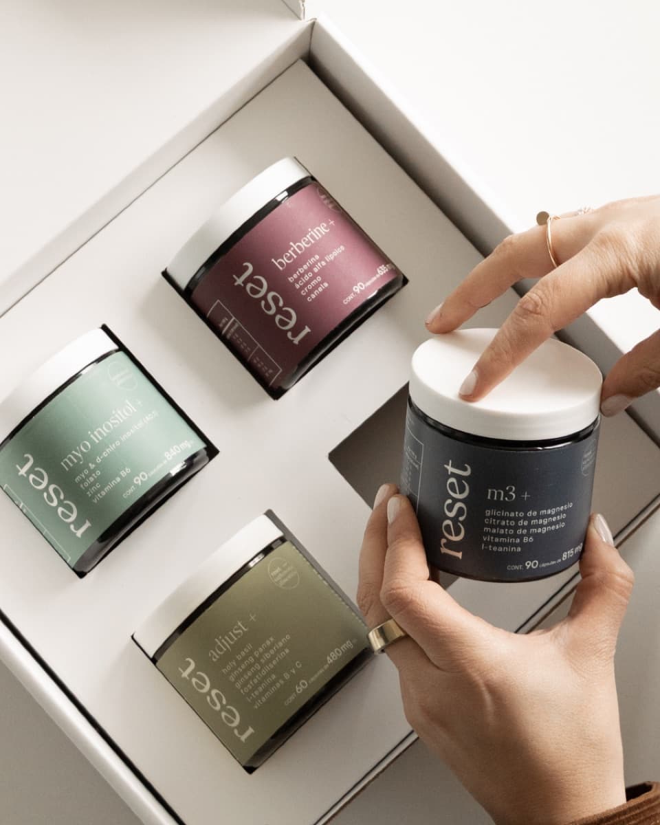 A hand placing a Reset Wellness m3+ magnesium jar into a gift box beside berberine+, myo inositol+ and adjust+ jars