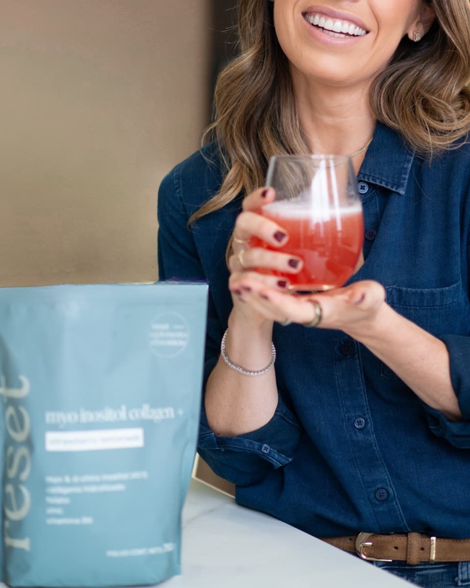 A woman holding a glass of strawberry lemonade myo inositol collagen+ next to the Reset Wellness pouch